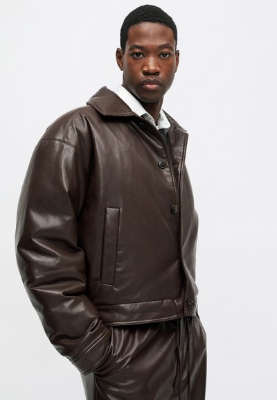 Moški v temno rjavi usnjeni jakni in ustreznih hlačah, stoji z eno roko v žepu ob enobarvni svetli ozadju.