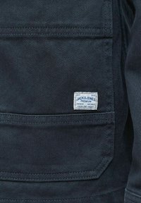 Tumši zilas auduma kabata ar baltu taisnstūra formālu etiķeti, uz kuras ir uzšūts teksts "Jack & Jones Premium Vintage Archive From The Vault".