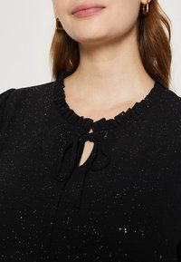 Blouse noire avec un col à volants, détail en forme de trou de serrure et fermeture à nouer. Présente un motif de paillettes subtil sur le tissu.