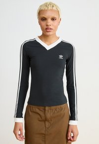 Camisola preta de manga longa com um decote em V branco e punhos brancos, apresentando riscas laterais brancas e um pequeno logótipo branco da Adidas no peito.