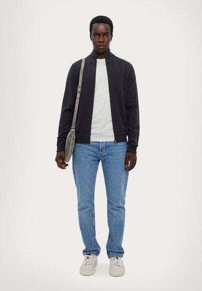 Dunkle Zip-Jacke über einem hellen T-Shirt, blaue Jeans und beigen Sneakern. Umhängetasche mit langem Riemen, einfarbiger Hintergrund.