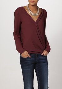 Top de manga larga burdeos con un diseño de escote en V cruzado, hecho de una tela suave, combinado con unos jeans de color azul oscuro y un collar plateado grueso.
