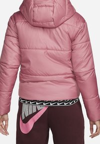 Rosa puffjacka med huva, med sömmar och en slät textur. Bärs över svarta och vita Nike-mönstrade träningsbyxor.