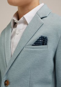 Svjetloplava jakna s teksturiranim završetkom, sa jednim dugmetom i mornarskoplavim uzorkovanim rupčićem u džepu, nosi se preko bijele košulje.