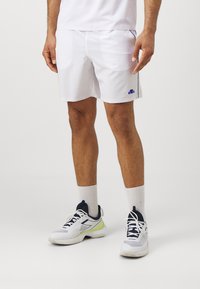 Weiße Sportshorts mit seitlichen marineblauen Akzenten, ausgestattet mit einem elastischen Bund und einem kleinen blauen Logo. Kombiniert mit weißen Turnschuhen und Knöchelsocken.
