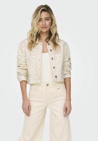 Veste croppée de couleur crème avec des motifs en œillets, fermeture à boutons et deux poches avant, assortie à un pantalon large.