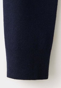 Gros plan d'une manche de pull en maille bleu marine avec un poignet côtelé sur un fond clair uni.