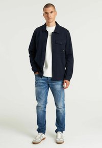 Marineblaues Hemd-Jacke mit Knopfverschluss, Fronttasche, kombiniert mit einem weißen T-Shirt, blauen Jeans und grauen Sneakers mit braunen Sohlen.