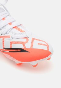 Botas de fútbol blancas con acentos naranjas, hechas de material sintético texturizado. Presenta un logotipo prominente y letras grandes en el lateral.