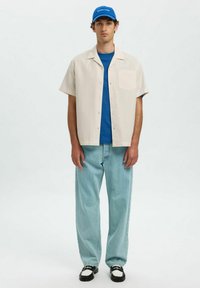 Lichtbeige kortarm shirt met ruitpatroon, gelaagd over een blauwe T-shirt, gecombineerd met lichtblauwe rechte jeans en zwarte instappers.