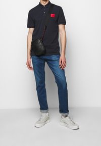 Polo bleu marine avec un patch logo rouge, associé à un jean bleu clair et des baskets grises. Sac bandoulière noir avec une surface texturée.