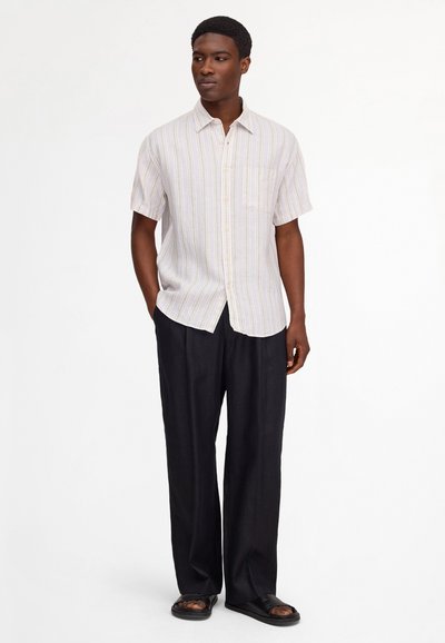 Man standing, wearing short-sleeve, light striped button-up shirt, black wide-leg pants, and black open-toe sandals on white background.