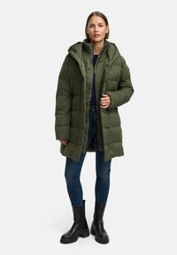 Zaļa polsterēta jaka ar kapuci, ar rāvējslēdzēju priekšā, pogu aizdari un teksturētu audumu. Kombinēta ar zilām džinsām un melniem masīviem zābakiem.