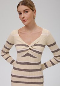 Robe longue à manches longues côtelée en beige avec des rayures marron foncé et rose clair, présentant un design à l'avant torsadé et un décolleté en V profond.