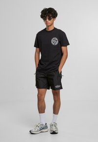 T-shirt noir avec un design graphique rond, shorts noirs avec un détail de texte, chaussettes blanches et chaussures de sport argentées. Modèle avec des cheveux bouclés et des lunettes de soleil.