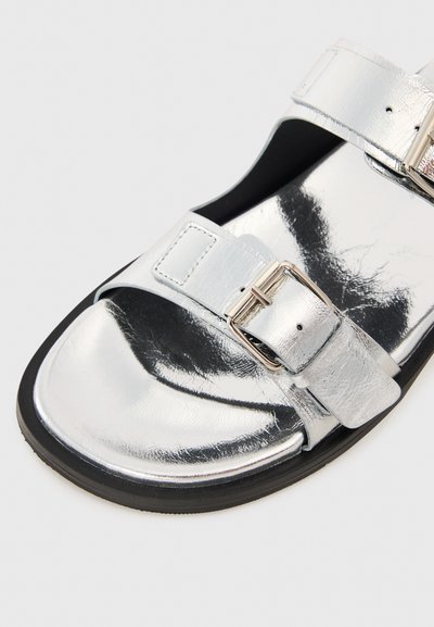 Sølv metallic sandaler med to justerbare stropper og spænde accenter. Har en glat, blank overflade på en sort gummisål.