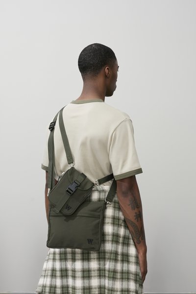 Sac bandoulière en polyester vert avec sangle réglable, fermeture à boucle, poche avant à rabat et détail logo, porté sur une chemise beige et une jupe à carreaux.