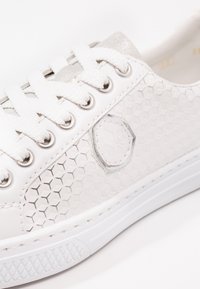 Baskets blanches avec motif hexagonal, lacets blancs, œillets argentés, et un logo octogonal embossé sur le panneau latéral.