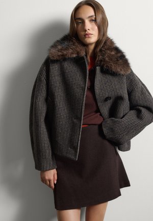 Junge Frau trägt eine dunkel karierte Jacke mit großem Fellkragen, ein braunes geripptes Oberteil und einen dunkelbraunen Rock, steht vor einer hellen Wand.