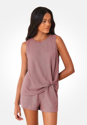 Mujer con cabello oscuro y largo, que lleva una camiseta sin mangas texturizada en color malva con un nudo torcido y shorts a juego, mirando hacia un lado.