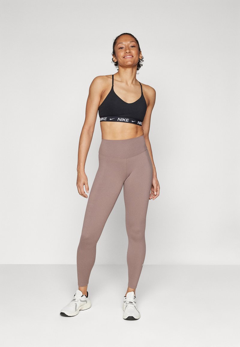 Zwarte sportbeha met wit logo, gecombineerd met lichtbruine high-waisted leggings. Model draagt witte sportschoenen. Eenvoudig, getailleerd ontwerp.