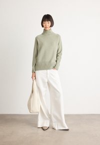 Woman in a sage green turtleneck sweater, wide white pants, beige pointed shoes, holding a cream tote bag, standing against a white wall.