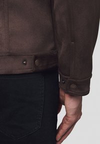 Brązowa skórzana kurtka z zamszu o fakturowanej powierzchni, z zapięciem na zatrzask i akcentowanymi mankietami, noszona na czarnych dżinsach.