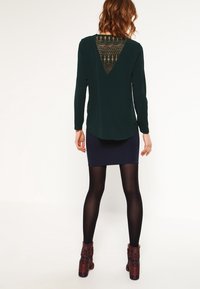 Haut vert foncé à manches longues avec un dos en dentelle décorative, associé à une jupe côtelée bleu marine. Le modèle porte des collants noirs et des bottines à motifs.