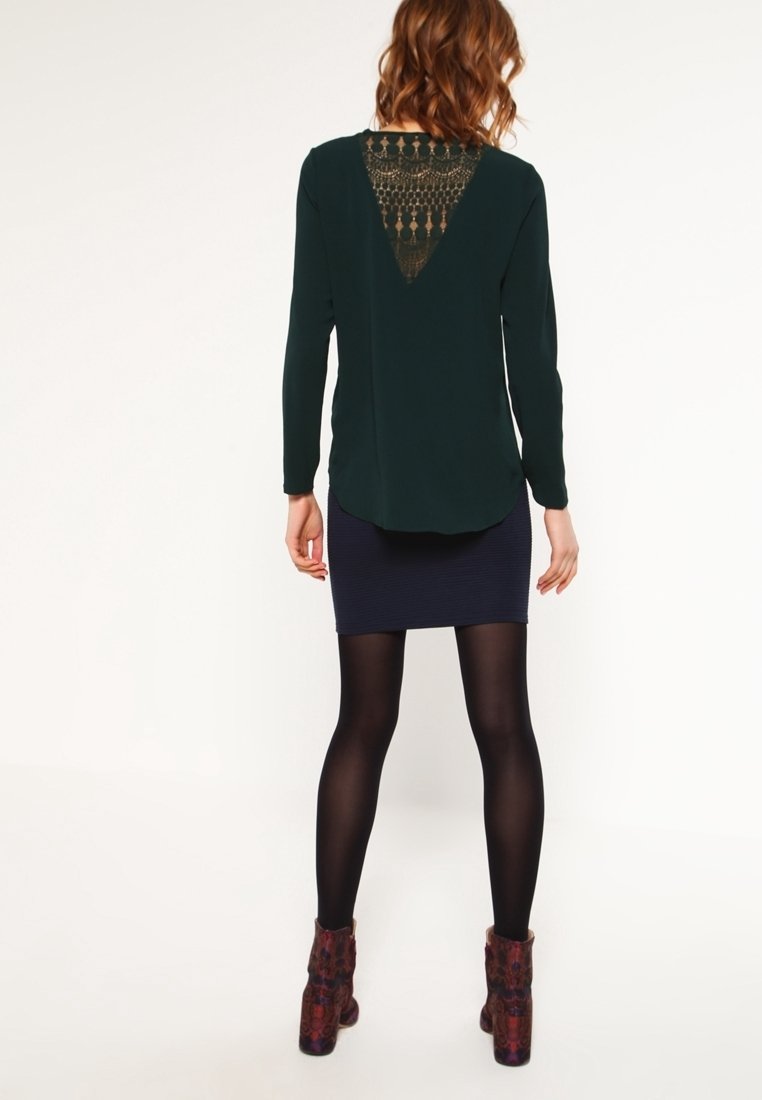 Haut vert foncé à manches longues avec un dos en dentelle décorative, associé à une jupe côtelée bleu marine. Le modèle porte des collants noirs et des bottines à motifs.