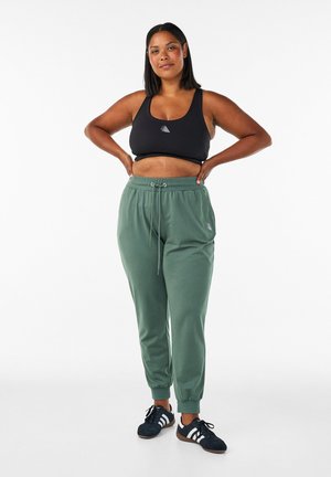 Selvsikker kvinde iført sort sports-bh, grønne joggingbukser og sorte sneakers står med hænderne på hoften mod en hvid baggrund.