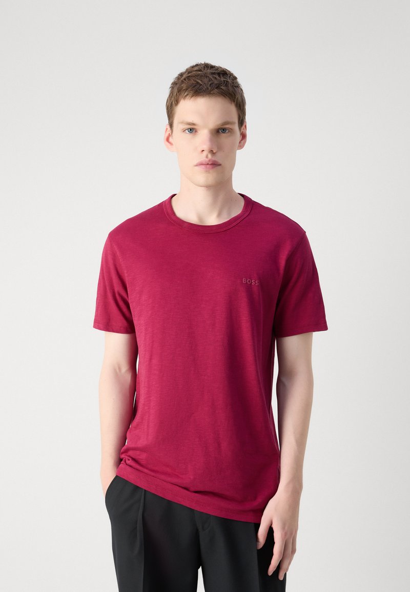 T-shirt rouge à col rond en tissu doux. Il présente des manches courtes et un petit logo sur la poitrine gauche. Porté avec un pantalon noir.