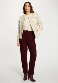 Donna in piedi con indosso un giubbotto crema a trama sopra un maglione chiaro, pantaloni bordeaux con cintura e stivali con tacco alla caviglia coordinati.