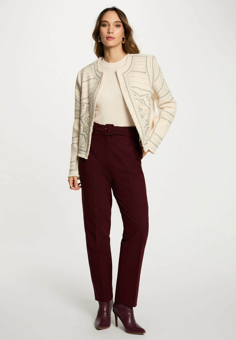 Donna in piedi con indosso un giubbotto crema a trama sopra un maglione chiaro, pantaloni bordeaux con cintura e stivali con tacco alla caviglia coordinati.