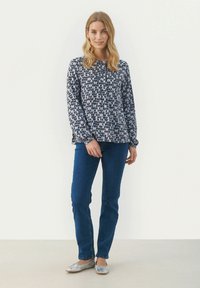 Blusa a maniche lunghe in navy con un motivo geometrico, abbinata a jeans blu e ballerine argento. La blusa presenta una chiusura frontale con bottoni e una vestibilità comoda.