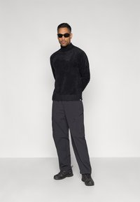 Schwarzer, strukturierter Rollkragenpullover, tailliert geschnitten, kombiniert mit schwarzen Cargo-Hosen mit Taschen und Reißverschlüssen, dazu schwarze Sneaker.