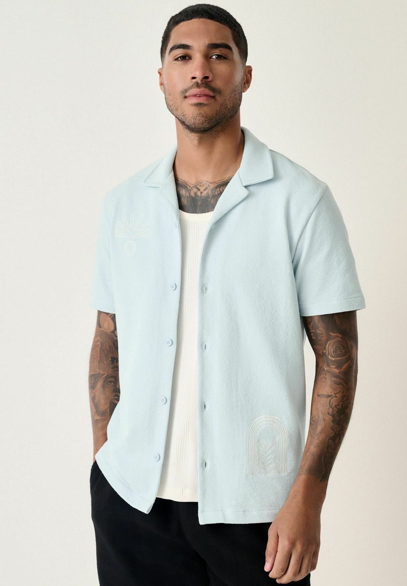 Camisa de manga corta de color azul claro, hecha de una tela texturizada, que presenta un cierre de botones en la parte delantera y sutiles diseños bordados en el pecho.