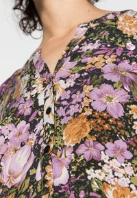 Blusa de padrão floral em preto, com flores roxas, laranjas e verdes. Decote em V notável e detalhes de botões. Tecido macio e texturizado.