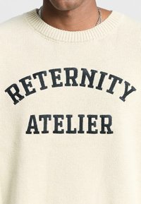 Cream knitted sweater featuring black embroidered text "RETERNITY ATELIER" in a curved layout. Round neckline and soft texture.