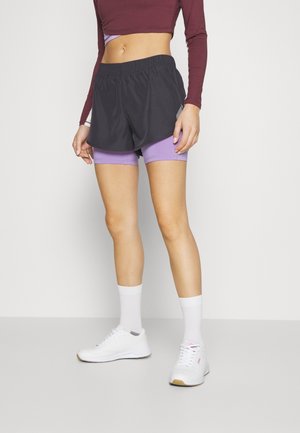 Frau trägt schwarze Shorts über lila Radlerhosen, ein bordeauxfarbenes langärmeliges Crop-Top, weiße Socken und weiße Sneakers und steht vor einem schlichten Hintergrund.