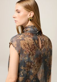 Bluse mit floralem Muster aus transparenter Stoff, mit einem Stehkragen, Knopfdetails am Rücken und umgeschlagenen Ärmeln. Die Farben umfassen Grau, Braun und Gold.