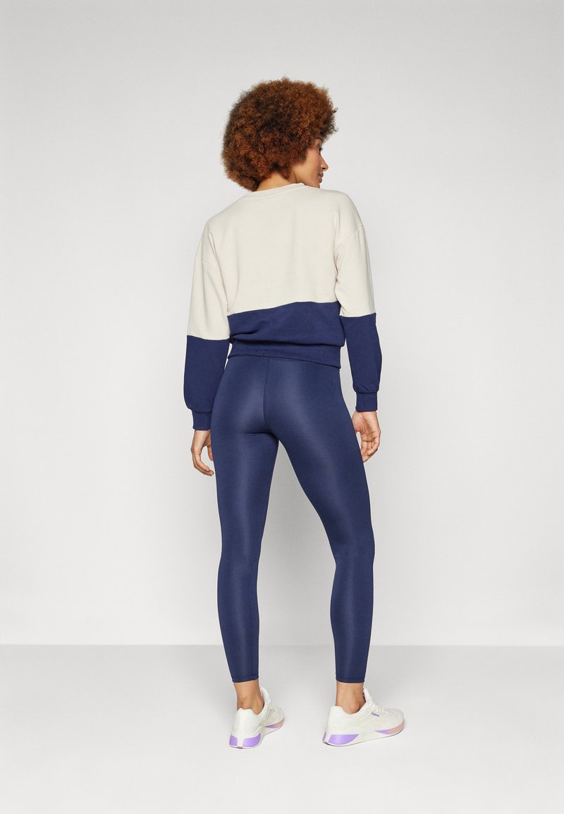 Kremowa i granatowa bluza z kolorowym blokiem, dopasowane granatowe legginsy. Model ma na sobie białe buty sportowe z purpurowymi akcentami.