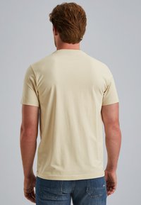 Camiseta de algodón beige claro con mangas cortas, textura suave y corte clásico. La vista trasera muestra un diseño simple y sin adornos.