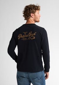 Zwart katoenen shirt met lange mouwen, voorzien van een tan kleurige grafische print op de achterkant met tekst "Petrol Ind & Co" en een arendontwerp. Blauwe denim jeans.