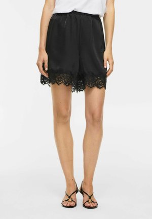 Vrouw draagt zwarte satijnen korte broek met kanten afwerking aan de zoom en zwarte hoge sandalen met bandjes, staand tegen een witte achtergrond.