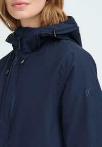Mornarsko modra vodoodporna jakna z visokim ovratnikom in nastavljivo kapuco. Ima sprednji zadrgo in subtilen logotip na rokavu. Gladka tekstura.