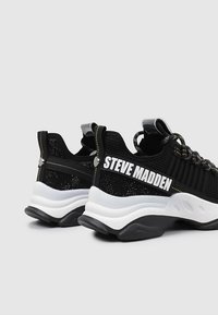Baskets en mesh noires avec des accents blancs, présentant une épaisse semelle noire et le logo "STEVE MADDEN". Comprend des lacets à motifs et des côtés texturés.