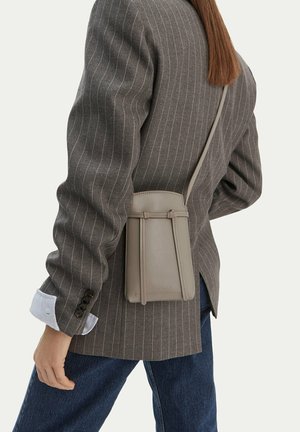 Osoba ubrana w szarą marynarkę w prążki i niebieskie dżinsy niesie małą beżową skórzaną torebkę na ramię na plecach.