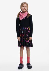 Ung pige med to fletninger, if ørt en sort langærmet bluse, farverig nederdel med stjernemønster, lyserødt tørklæde, lyserøde sokker og sorte sko, stående.