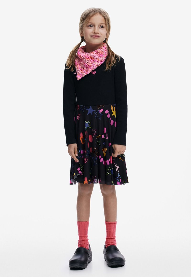 Ung pige med to fletninger, iført en sort langærmet bluse, farverig nederdel med stjernemønster, lyserødt tørklæde, lyserøde sokker og sorte sko, stående.