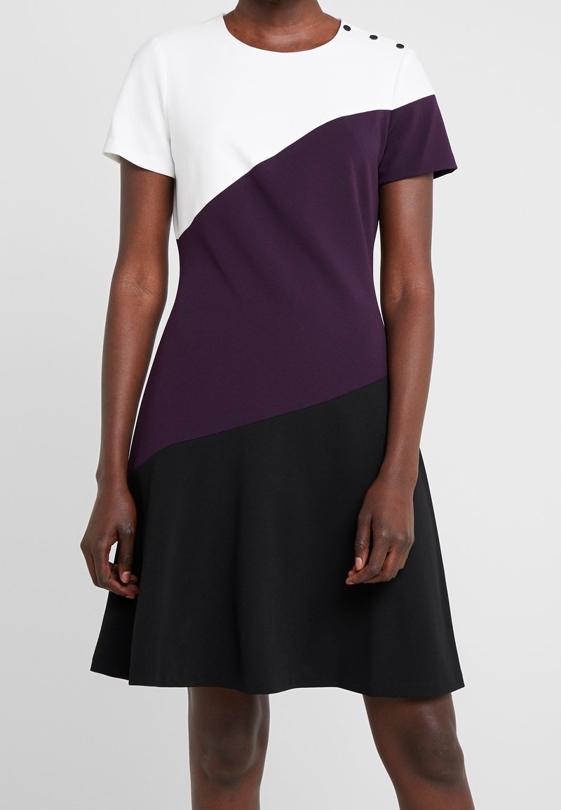 Robe à manches courtes, longueur genou, avec haut blanc, milieu violet, jupe noire et trois boutons sur l'épaule gauche.